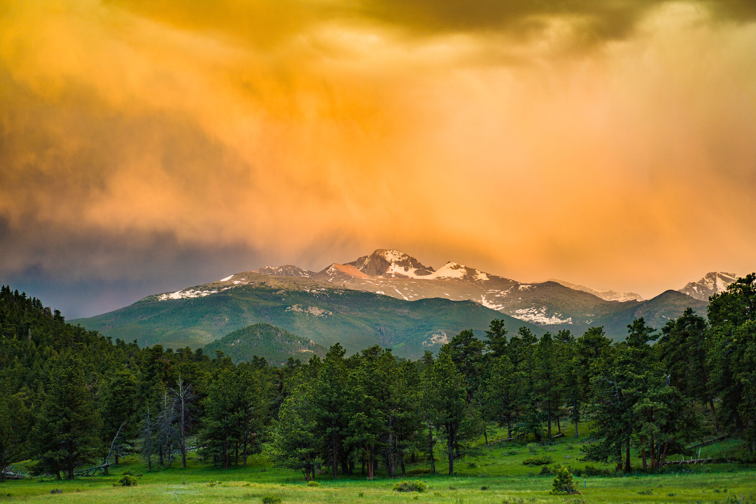 A mountain range with snow-capped peaks rises above a dense green forest, while dramatic orange and yellow clouds fill the sky during sunset or sunrise.