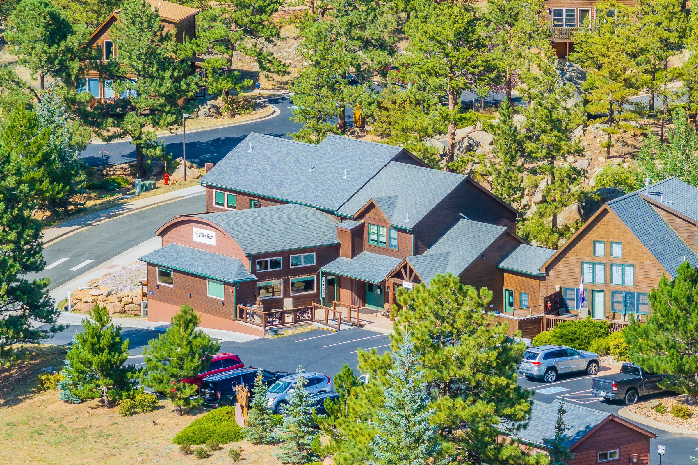 A brown building with a sign reading "Office" is surrounded by trees and other wooden cabins. There is a parking lot with several cars in front and a winding road behind the buildings.